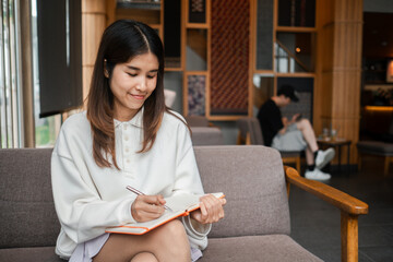 A young woman sits comfortably in a cafe, writing in her notebook, surrounded by a modern and cozy interior.