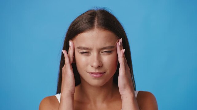 A young Caucasian woman with long hair is shown with a pained expression, gently touching her temple as if feeling migraine or a headache. The vibrant blue backdrop enhances her focused demeanor.