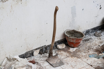 A hoe stands in front of a white wall photographed from the left corner, with a red bucket and some debris and dirt from the building around it