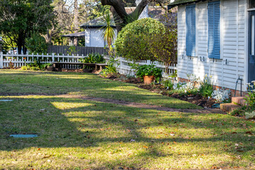 Suburban front yard with a white picket fence, trimmed lawn, flower beds, and an old weatherboard house. A peaceful residential garden in a traditional Australian neighborhood.