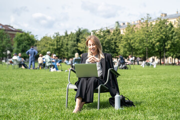 Laughing mature woman watching movie or video call on laptop in park on summer break. Female online entertainment outdoors, remote connectivity, leisure time, flexible work, urban park relax