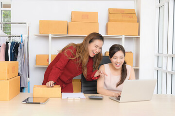 Two smiling Asian women, business partners, happily discuss operations and review documents for their online store in a bright home office setting.