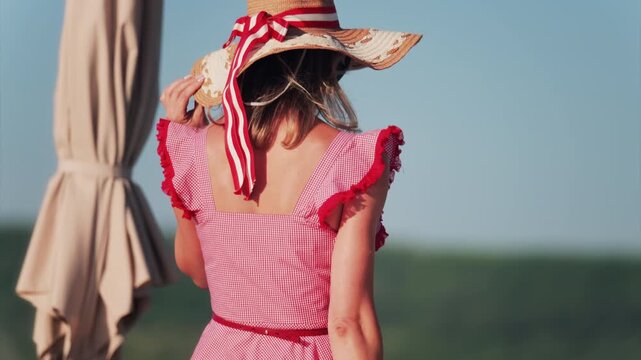 Woman in a red gingham dress and wide hat with a ribbon walking on a terrace