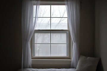 A softly lit interior scene featuring a tall multi paned window draped with sheer white curtains revealing a muted outdoor view