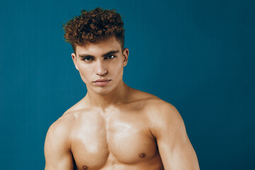 Confident young man with curly hair posing shirtless against a solid blue background, expressing...
