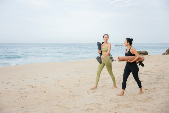 Two women walking on the beach carrying yoga mats after yoga class