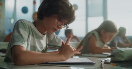 Primary School Boy Writing School Test, Doing Task in Notebook. Female Teacher Walking Between Desks, Controlling Group of Diverse Kids During the Exam Process in Modern Classroom. Close Up Shot. - Powered by Adobe