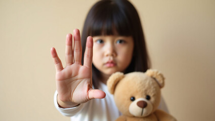 A serious young child holding a teddy bear with her hand held up in a stop gesture. The child is looking directly at the viewer with a stern expression. The teddy bear is clutched in her other arm.