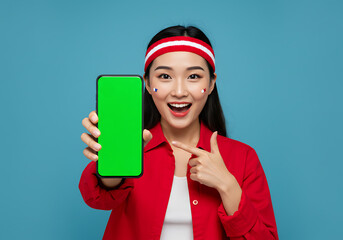 Cheerful Asian woman supporting her national team, showing a mobile phone with a blank chroma key screen for mockups on a blue background.