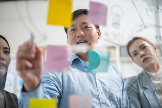 Asian business team in a creative meeting, collaborating and sharing ideas using colorful notes on a glass wall. A modern teamwork setting showing brainstorming, innovation, and planning in office
