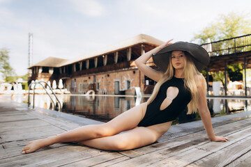 Fashion model relaxing by the poolside wearing a black swimsuit and wide-brimmed hat