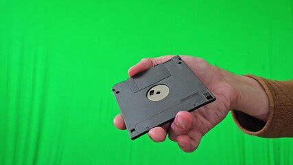A nostalgic and technical mood is captured as a hand holds a classic 3.5-inch floppy disk, ready for integration into various digital or historical themes against a vibrant green screen - Powered by Adobe