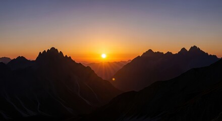 Dramatic mountain peaks emerge from the darkness as the sun sets casting a warm orange glow silhouette