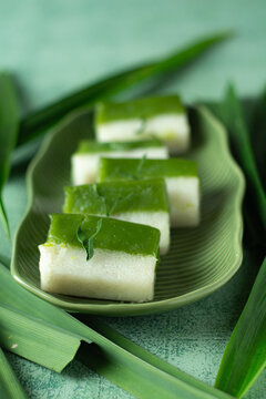 Traditional Indonesian Kue Talam Pandan on Green Plate, A close-up shot of traditional Indonesian dessert Kue Talam Pandan, neatly arranged on a green ceramic plate with pandan leaves as background. T