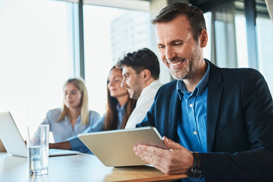 Happy mature businessman using tablet PC with colleagues working in background