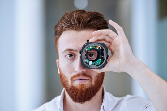 Young man looking through open camera lens at office