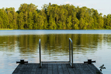 A wooden dock with a swimming ladder extending into a calm lake surrounded by lush green forest. Peaceful lakeside pier with metal handrails reflecting in tranquil water