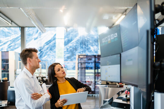 Businesswoman examining computer monitors through tablet PC standing by businessman with arms crossed