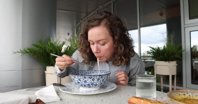 Lady eats noodles from soup with chopsticks. Cute young woman savoring a traditional dish at a stylish outdoor caf, surrounded by greenery, showcasing a moment of cultural dining