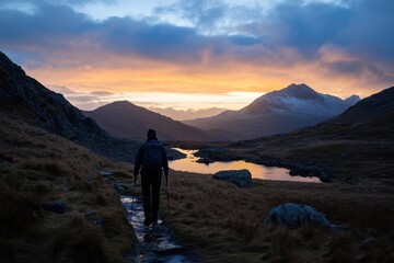 Obraz premium Hiker treks along mountain path at sunset with reflections on still water in the background, Silhouette of hiker trekking mountain at sunset Snowdonia