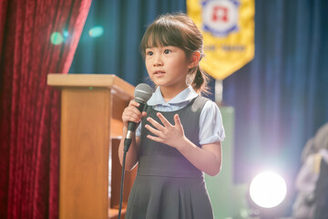 Young Girl Passionately Delivers Speech on Stage at School Event in Bright Setting