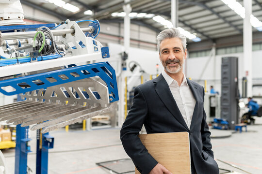 Happy businessman holding laptop standing by machinery in factory
