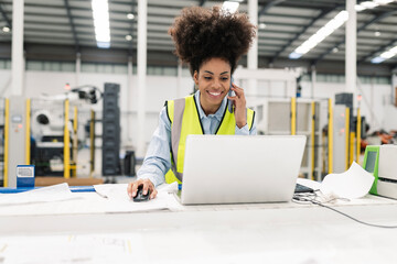 Smiling engineer working on laptop talking on mobile phone in factory