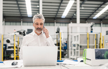 Businessman talking on smart phone standing with laptop at desk in factory