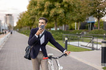 Businessman talking on speaker phone wheeling with bicycle on footpath