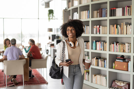 Smiling businesswoman with backpack holding smart phone walking in office