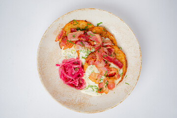 A plate features three golden pancakes with meats, pickled onions, and a sauce drizzle