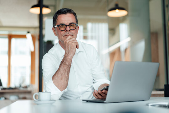 Confident businessman with hand on chin sitting with laptop on table at office