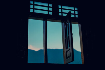 Dramatic dark window frame with an open shutter reveals a serene blue sky and distant mountain silhouettes.