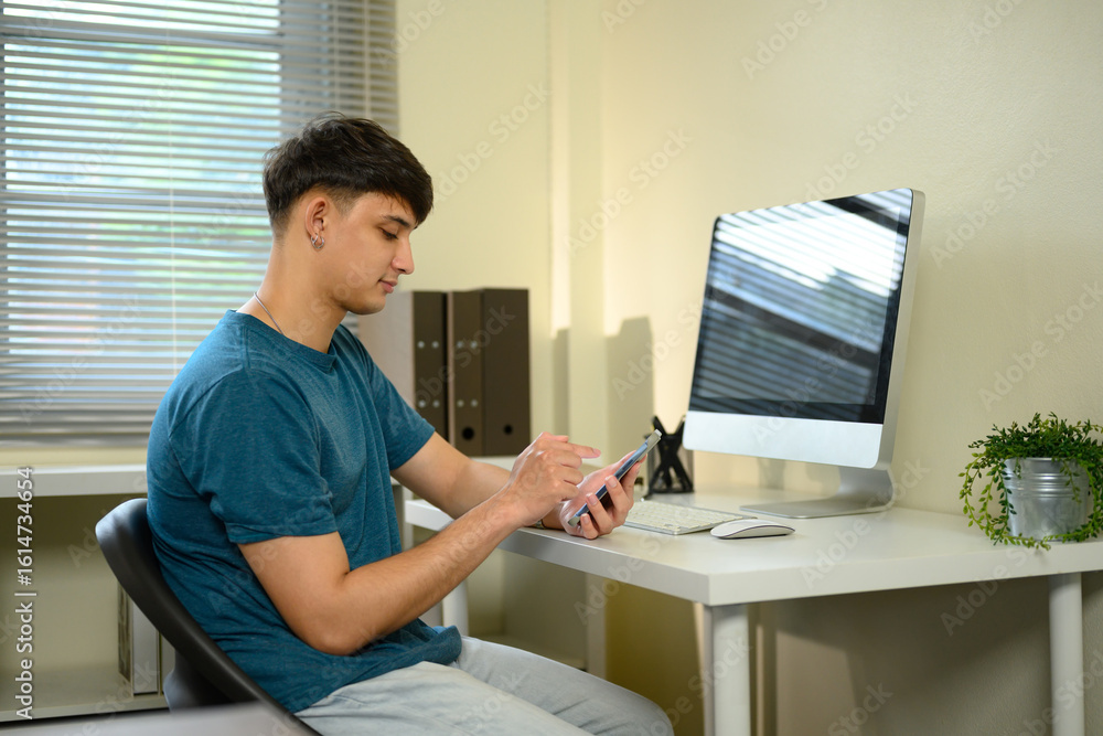 Aufkleber young man sitting at desk using smartphone in front of desktop computer in a home office