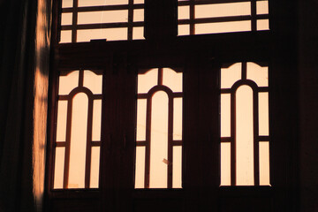 Golden sunset light streams through classic wooden windows with intricate patterned glass, creating warm indoor ambiance.