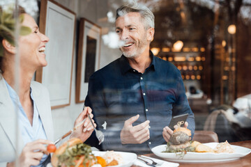 Happy businesswoman with businessman holding mobile phone having lunch in cafe seen through glass