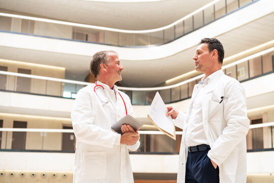Doctor with hand in pocket discussing with colleague holding tablet PC in hospital
