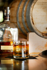 A glass of whiskey on a bar with the bottle and a wood barrel