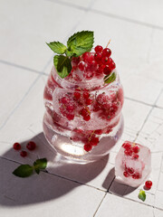 Iced drink with redcurrant frozen in ice cubes garnished with mint leaves on a cracked white tile....