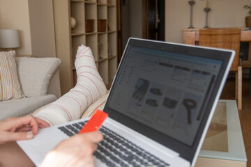 Person with broken leg resting on sofa at home, shopping online using laptop and credit card,...