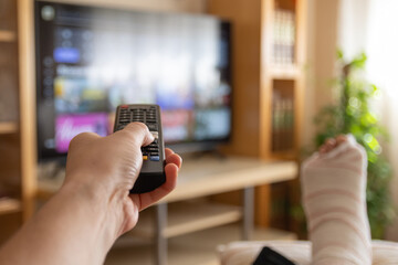 Person with a broken leg wrapped in a cast enjoys streaming services from home, using a remote...