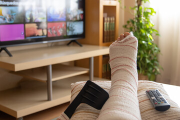 Person with a broken leg wrapped in a cast rests their foot on a pillow, watching streaming...
