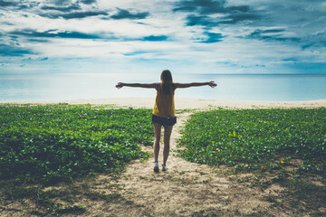 Young woman with open arms facing the sea under dramatic cloudy sky. Wild beach, green plants and...