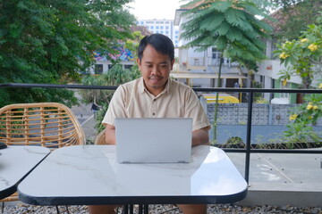 Smiling man uses a laptop at an outdoor cafe table, surrounded by greenery and urban buildings,...