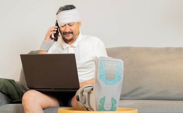 Happy smiling young guy with broken leg and head sitting in sofa at home, using mobile phone and laptop.