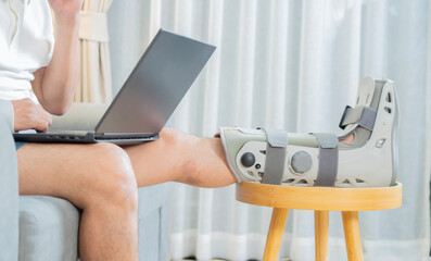 Side view shot of man with broken leg working on laptop at home sitting on sofa