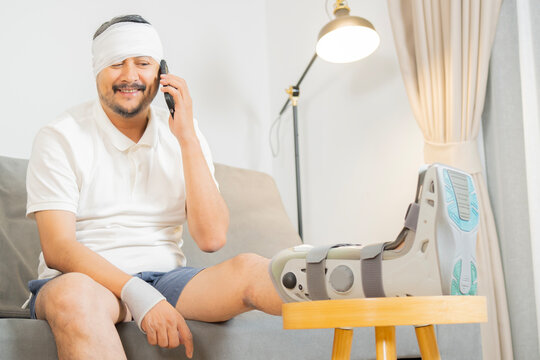 Sick adult Asain man sit on sofa wearing bandages his head, wrist and leg using smartphone