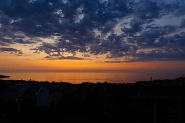 Sunset over the sea and cityscape. Orange and purple light in the sky, dramatic clouds, calm reflections on water – summer evening view from above, peaceful and atmospheric.