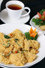 A close-up shot of a plate filled with golden-fried crispy shrimp coated in salted egg yolk floss, garnished with curry leaves, chili, and a carrot flower