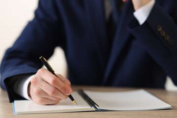Man writing in notepad with pen at wooden table against light background, closeup